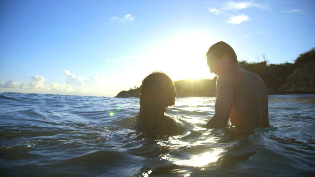 Man and woman in the ocean smiling while the sun sets over the hill